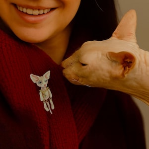 May include: A white cat brooch with blue eyes and a gold chain is pinned to a red garment. A hairless cat is nuzzling the garment. The brooch is a miniature sculpture of a cat.