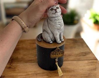 May include: A decorative black container with a wooden top, featuring a gray cat figurine. The container has a gold-colored tag that reads "BELLA" and a gold tassel. The cat is sitting upright.