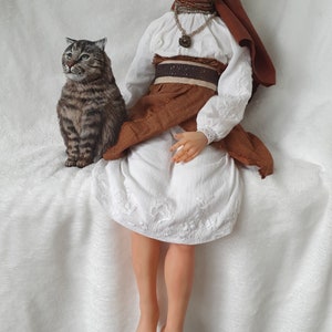 May include: A woman wearing a white dress with brown embroidery and a brown head scarf sits on a white fluffy surface. She is wearing a brown belt with a gold buckle and a necklace with a pendant. A tabby cat sits beside her.