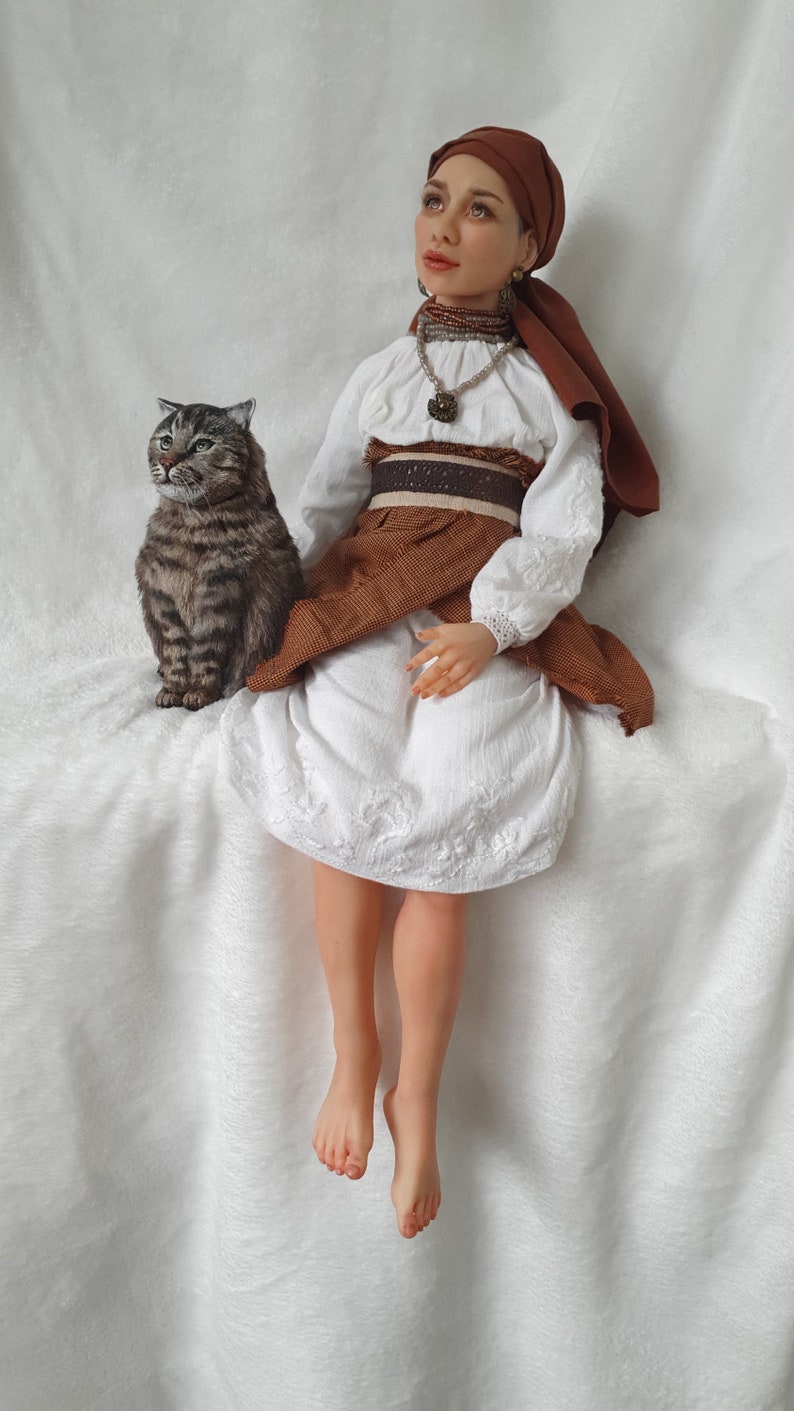 May include: A woman wearing a white dress with brown embroidery and a brown head scarf sits on a white fluffy surface. She is wearing a brown belt with a gold buckle and a necklace with a pendant. A tabby cat sits beside her.