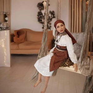 May include: A woman wearing a white embroidered blouse with a brown skirt and a red headscarf sits on a rope swing. She is wearing silver jewelry and has a neutral expression.