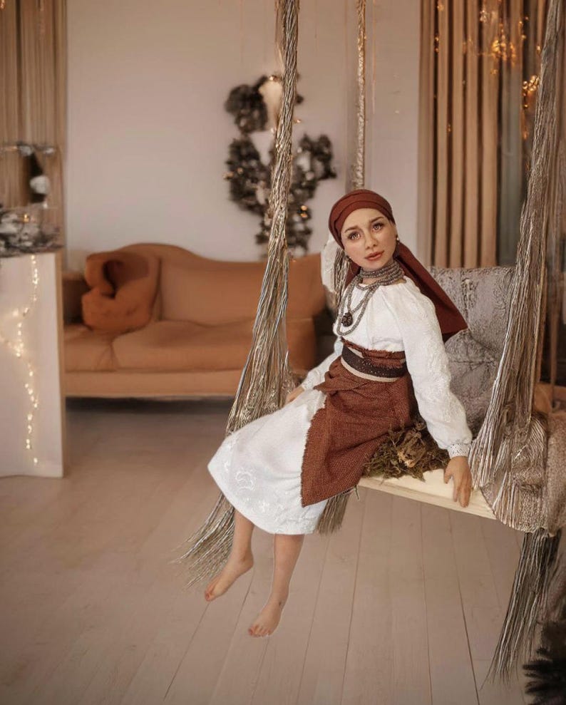 May include: A woman wearing a white embroidered blouse with a brown skirt and a red headscarf sits on a rope swing. She is wearing silver jewelry and has a neutral expression.