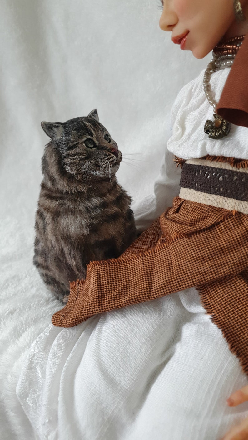 May include: A brown and gray tabby cat sits on a brown and white fabric, looking up at a doll with a white dress and brown skirt. The doll has a white pearl necklace and a brown leather belt.