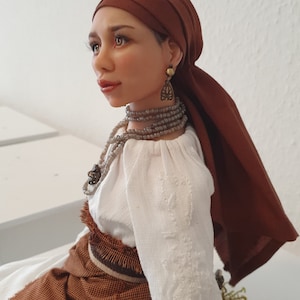 May include: A handcrafted doll with brown hair and a brown headscarf. She is wearing a white blouse with embroidery, a brown skirt, and a beaded necklace. The doll is sitting on a piece of wood with moss.