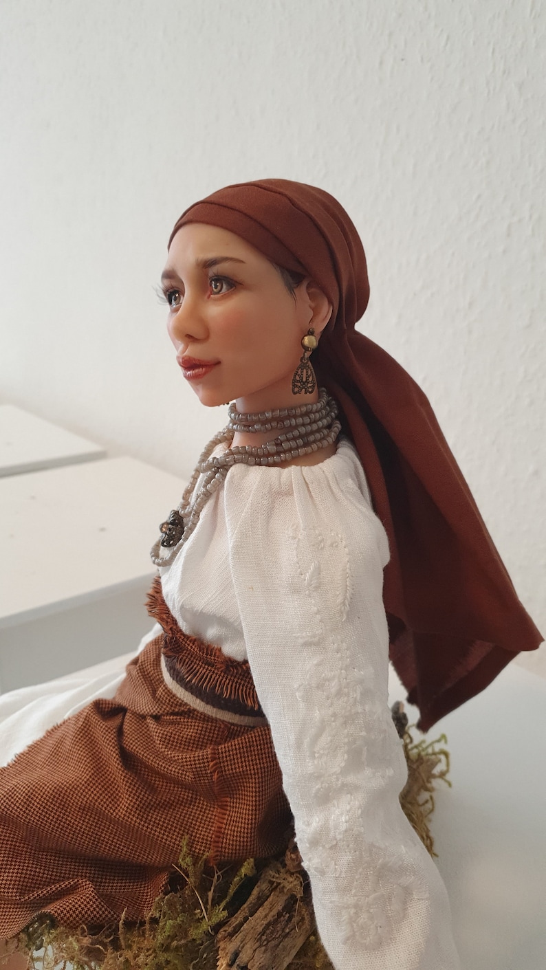 May include: A handcrafted doll with brown hair and a brown headscarf. She is wearing a white blouse with embroidery, a brown skirt, and a beaded necklace. The doll is sitting on a piece of wood with moss.