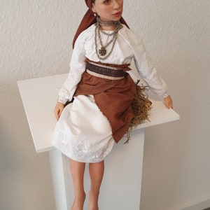 May include: A porcelain doll wearing a white dress with brown embroidery, a brown head scarf, and a brown skirt. The doll is sitting on a white pedestal.