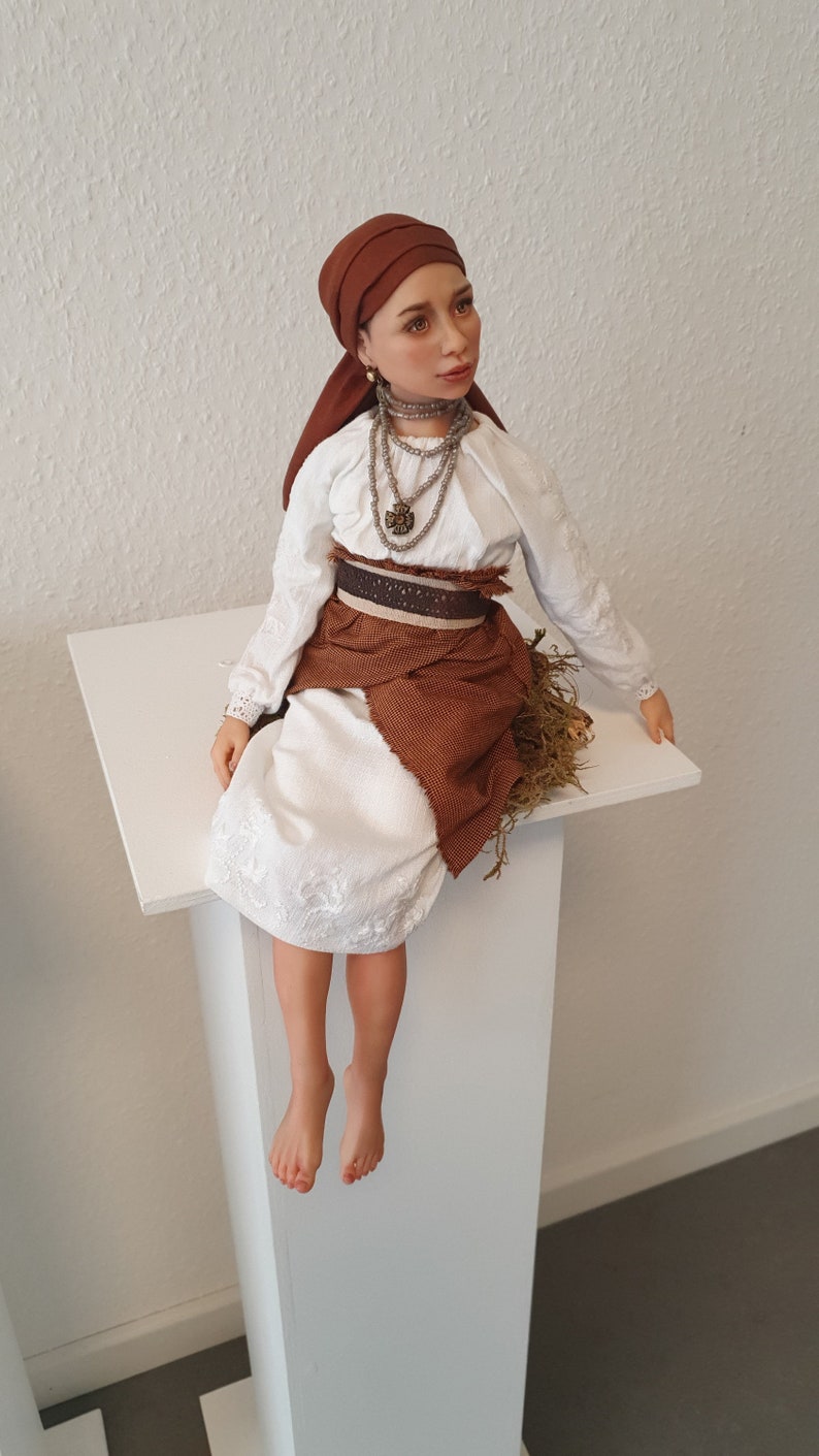 May include: A porcelain doll wearing a white dress with brown embroidery, a brown head scarf, and a brown skirt. The doll is sitting on a white pedestal.