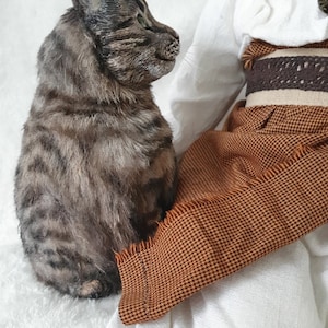 May include: A brown and gray tabby cat figurine sitting on a white surface, looking to the right. A doll with a white dress and brown skirt is sitting in the background.