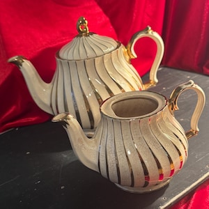 May include: A cream-colored teapot and creamer set with gold accents. The teapot has a lid and a curved handle, and both pieces feature vertical gold stripes. The set is on a dark surface with a red background.