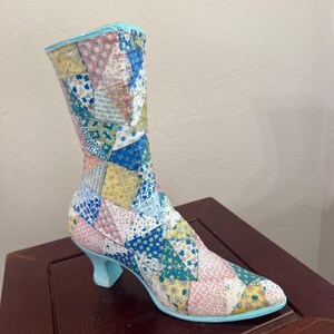 May include: A decorative boot-shaped object featuring a patchwork design of various triangular fabric patterns in shades of blue, yellow, pink, and white. The boot has a light blue trim along the top and base, and a small, curved light blue heel.