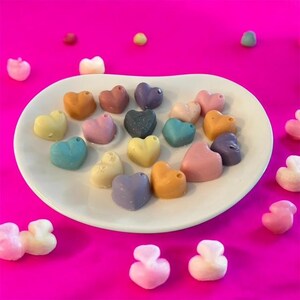May include: A white oval plate displays a selection of heart-shaped sweets in shades of yellow, orange, pink, blue, and purple. The candies are arranged on a vibrant pink surface, with a few more scattered around the plate.
