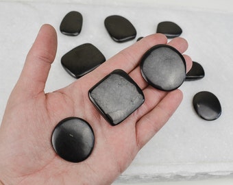 May include: A collection of black obsidian stones in various shapes, including round, square, and rectangular, displayed on a white surface.