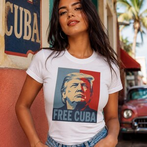 May include: A white short-sleeved t-shirt featuring a graphic print of Donald Trump in a stylized blue and red color block design. The text "FREE CUBA" is printed in bold blue capital letters at the bottom of the rectangular graphic on the chest.