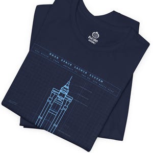 Op de afbeelding: Marineblauw t-shirt met een lichtblauw ontwerp van het NASA Space Launch System. De afbeelding bevat technische details en tekst. Het t-shirt is opgevouwen, met een ronde hals. Het merklogo is rechtsboven zichtbaar.
