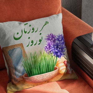 Puede incluir: Una almohada blanca con un diseño verde y blanco que presenta una maceta de hierba verde, una flor morada y el texto "Nowruz Mubarak".