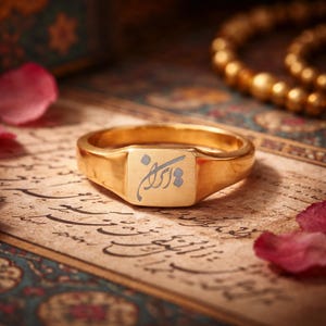 Puede incluir: Un anillo de sello dorado con un diseño grabado en una cara cuadrada. El anillo está colocado sobre una superficie con caligrafía y rodeado de pétalos de rosa y una pulsera de cuentas doradas. El diseño del anillo está en una fuente de escritura.
