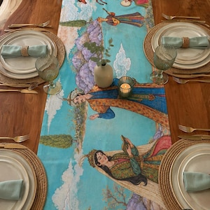May include: A long rectangular table runner featuring a vibrant illustration of figures in traditional attire, horses, and stylized clouds on a light blue background. The runner is centered on a wooden table with four place settings, each including a plate, a light green napkin with a woven ring, a glass, and gold-toned cutlery.