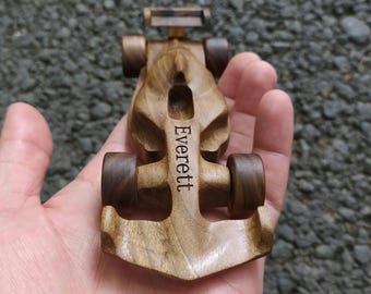 Puede incluir: Un pequeño coche de carreras de juguete de madera con un acabado suave y pulido. El coche presenta un diseño aerodinámico de cabina abierta con cuatro ruedas de madera oscura. El nombre Everett está grabado en una fuente oscura en el centro.