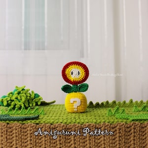 Pode incluir: Amigurumi de Flor de Fogo de crochê, com uma cabeça de flor vermelha e amarela, folhas verdes e um corpo amarelo com uma interrogação branca. A cena inclui grama verde de crochê e o texto "Amigurumi Pattern".