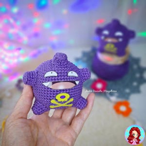 May include: A handmade purple crochet toy with a smiling face, white teeth, and a yellow skull and crossbones emblem. The toy is held in a hand, with a blurred background featuring another similar toy and colorful lights.