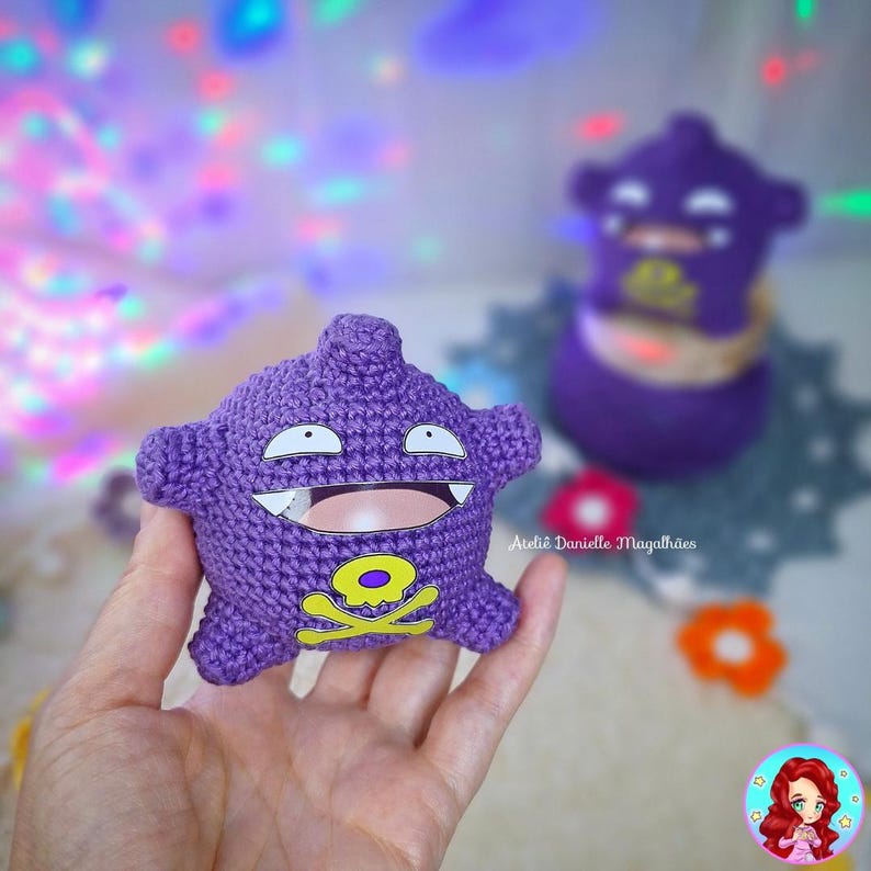 May include: A handmade purple crochet toy with a smiling face, white teeth, and a yellow skull and crossbones emblem. The toy is held in a hand, with a blurred background featuring another similar toy and colorful lights.