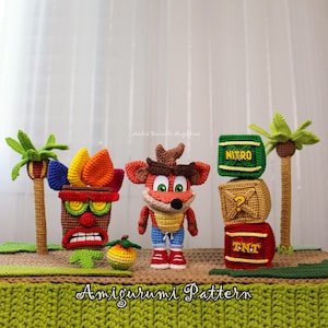 Pode incluir: Uma coleção de figuras amigurumi de crochê, incluindo um personagem marrom com grandes olhos verdes, uma máscara marrom com penas coloridas, uma fruta amarela, duas palmeiras e uma pilha de três cubos rotulados NITRO, um ponto de interrogação e TNT.