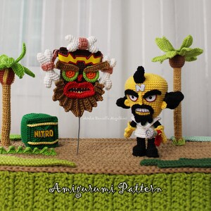May include: A crochet display featuring a yellow character with a black mohawk and beard, a brown mask with white bones, a green Nitro crate, and two palm trees on a textured base. The text Amigurumi Pattern is printed at the bottom of the image.