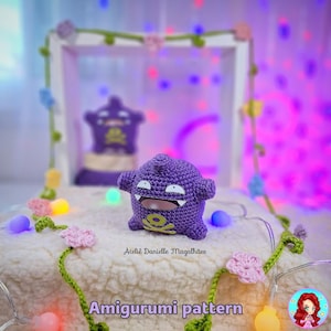 Op de afbeelding: Een handgemaakt paars gehaakt amigurumi-speeltje met een lachend gezicht en een geel schedel- en kruisbotontwerp. De tekst "Amigurumi pattern" is onderaan zichtbaar. De achtergrond omvat een decoratieve display met een vergelijkbaar speeltje en lichtslingers.