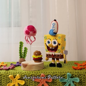 May include: Crocheted SpongeBob SquarePants figure with a spatula, a rocket, and a burger, alongside a pink jellyfish and green seaweed. The scene is set on a green crocheted surface with starfish accents. The text "Amigurumi Pattern" is visible.