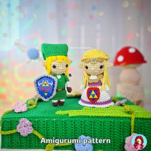 Pode incluir: Figuras amigurumi de crochê de Link e Zelda de The Legend of Zelda. Link está com uma túnica verde com um escudo, e Zelda usa um vestido branco e roxo. As figuras estão em uma base verde com flores. Texto: Amigurumi pattern.