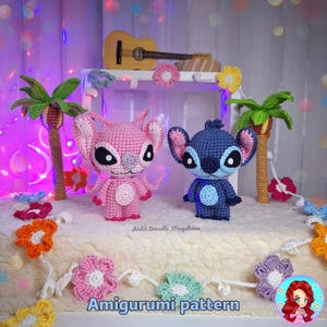 Pode incluir: Dois brinquedos de pelúcia amigurumi de crochê, um rosa e outro azul, com olhos grandes e detalhes costurados. Os brinquedos estão sentados em uma superfície branca com uma guirlanda de flores de crochê. O texto "Amigurumi pattern" é visível na parte inferior da imagem.