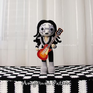 Può includere: Una bambola all'uncinetto con capelli neri, viso bianco con trucco nero a forma di stella e un vestito nero. La bambola tiene in mano una piccola chitarra elettrica piatta arancione e rossa. Il testo Amigurumi Pattern è visibile in basso.