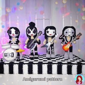 Pode incluir: Quatro bonecas de crochê feitas à mão que representam uma banda de rock, cada uma com pintura facial e roupa distintas. Uma boneca toca bateria com a palavra "ROCK". As outras três bonecas seguram guitarras. As bonecas estão em uma superfície xadrez em preto e branco.