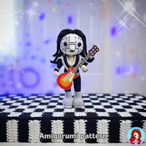 Pode incluir: Boneca amigurumi feita à mão de uma estrela do rock, vestindo uma roupa preta e prateada e segurando uma guitarra vermelha e laranja. A boneca tem um rosto branco com detalhes em preto e cabelo longo preto. Em uma superfície quadriculada em preto e branco. Texto: "Amigurumi pattern".