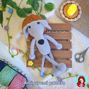 May include: A gray crocheted amigurumi dog toy with black eyes and nose, lying on a wooden crate. The dog is on a white fluffy surface with a green vine with colorful flowers. The text "Amigurumi pattern" is at the bottom of the image.