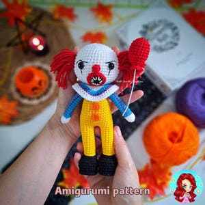 Pode incluir: Padrão de amigurumi de crochê para um palhaço assustador com um balão vermelho. A boneca está vestindo uma roupa branca e amarela com detalhes azuis. A boneca tem um nariz vermelho, olhos pretos e um sorriso assustador.