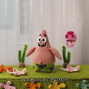 Könnte beinhalten: Eine gehäkelte Patrick Star Amigurumi-Puppe mit einem lächelnden Gesichtsausdruck, die grüne Shorts mit lila Akzenten trägt. Die Szene umfasst gehäkelte Seesterne, eine Qualle und Seetang. Der Text "Amigurumi Pattern" ist sichtbar.