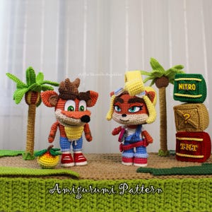 Pode incluir: Duas figuras amigurumi de crochê que lembram Crash e Coco Bandicoot, em pé sobre uma base marrom com duas palmeiras, uma fruta amarela e três caixas empilhadas com os rótulos NITRO, um ponto de interrogação e TNT. O texto Amigurumi Pattern é visível.