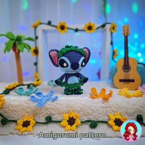 May include: A handmade crochet amigurumi of a blue alien character wearing a green lei and grass skirt. The scene includes a crochet palm tree, sunflowers, and a ukulele. The words "Amigurumi pattern" are at the bottom.