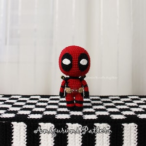 Könnte beinhalten: Handgefertigte Amigurumi-Puppe von Deadpool in Rot und Schwarz. Die Puppe hat ein detailliertes Design mit Gürtel, Trägern und einer Maske mit weißen Augen. Die Basis ist ein schwarz-weißes Schachbrettmuster. Der Text "Amigurumi Pattern" ist sichtbar.