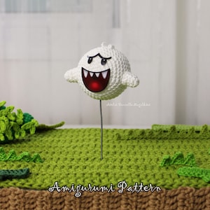Pode incluir: Um personagem Boo branco de crochê da série de videogames Super Mario. O fantasma amigurumi tem um sorriso largo e dentuço com um interior vermelho e olhos pretos. A figura é presa a um fio e colocada em um cenário de crochê verde. As palavras "Amigurumi Pattern" estão na parte inferior.
