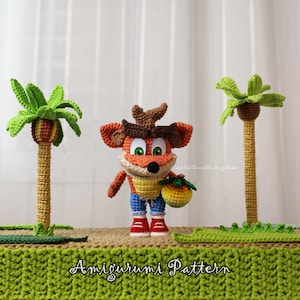 Pode incluir: Figura amigurumi de Crash Bandicoot de crochê com detalhes em laranja, marrom e amarelo, usando shorts azuis e sapatos vermelhos, segurando uma fruta amarela. Duas palmeiras de crochê ladeiam a figura. A base diz "Amigurumi Pattern".