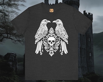 May include: Dark gray t-shirt featuring a white graphic of two ravens flanking a skull and ornate design. The ravens face each other, with detailed feather patterns. The design is centered on the shirt, creating a gothic or fantasy theme.