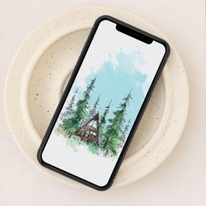 May include: A smartphone displays a watercolor illustration of a cabin nestled among tall green pine trees under a light blue sky. The phone rests on a speckled, off-white plate, which is placed on a white book.