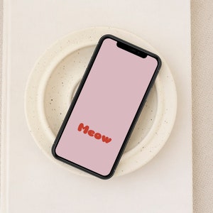 May include: A black smartphone with a pink screen displaying the word "Meow" in red bubble letters. The phone is resting on a white ceramic plate with a speckled pattern.