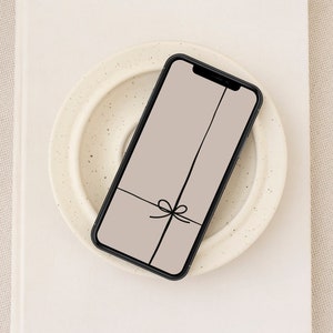 Könnte beinhalten: Ein schwarzes Smartphone mit einem hellbraunen Hintergrund auf dem Bildschirm. Der Hintergrund hat eine schwarze Linie in der Mitte und eine schwarze Schleife am unteren Ende der Linie.