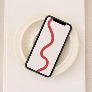 Könnte beinhalten: Ein schwarzes Smartphone mit weißem Hintergrund und einem roten abstrakten Linienmuster auf dem Bildschirm. Das Telefon liegt auf einem weißen Teller mit einem gesprenkelten Muster.