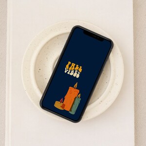 May include: A phone screen displaying a dark blue background with the text "Fall Vibes" in a retro font. Three candles are depicted, one orange, one green, and one brown, with flames.