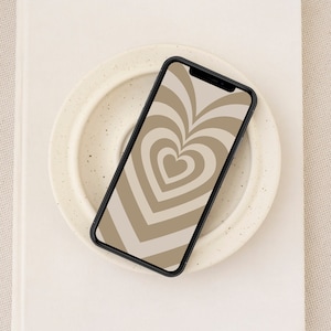 May include: A black smartphone with a brown and white heart pattern on the screen. The phone is lying on a white plate with a speckled surface.