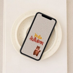 May include: A smartphone with a white background and a brown bear wearing a scarf. The text "Hello Autumn" is written in red and yellow on the screen.